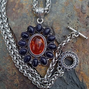May include: Silver-toned necklace featuring a large pendant with a central oval orange stone surrounded by dark blue stones. The pendant is attached to a thick chain with a toggle clasp. The necklace is displayed on a textured, dark surface.
