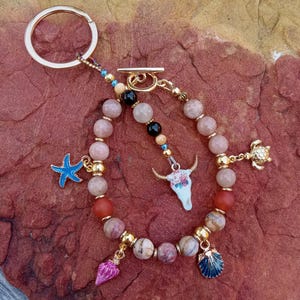 May include: A beaded keychain with a gold ring and clasp. The keychain features a strand of pink and red beads, accented with gold spacers and charms, including a blue starfish, a pink shell, and a turquoise cow skull.