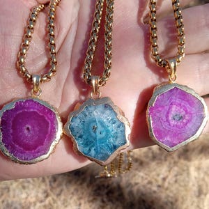May include: Three gold-tone necklaces with pendants. The pendants are slices of agate in shades of purple and blue, with gold-colored edges. The necklaces are displayed against a blurred background.