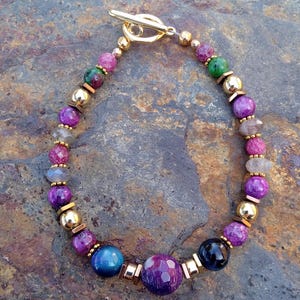 May include: A beaded bracelet featuring a toggle clasp and a mix of colorful beads. The bracelet includes purple, pink, green, and gold-colored beads. The bracelet is displayed on a textured, stone-like surface.