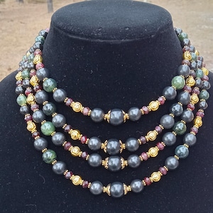 May include: A multi-strand necklace with gray, green, and burgundy beads. The necklace features large gray beads, smaller green and burgundy beads, and gold-colored accents. The necklace is displayed on a black mannequin.