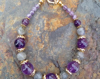 May include: A beaded bracelet featuring amethyst and gray labradorite stones. The bracelet has gold-colored accents and a toggle clasp. The amethyst beads are a deep purple, and the labradorite beads have a gray, iridescent sheen. The bracelet is displayed on a textured, stone surface.
