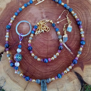 May include: A collection of gemstone jewelry including a necklace, bracelet, and earrings. The necklace features a large, rectangular pendant and is adorned with blue, purple, and gold beads. The bracelet and earrings complement the necklace with similar beadwork and gold accents.
