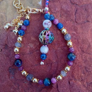May include: A beaded bracelet featuring a mix of blue, purple, and white stone beads interspersed with gold-toned metal beads. A central dangling charm consists of a white bead, a metallic iridescent filigree sphere, and a blue bead.
