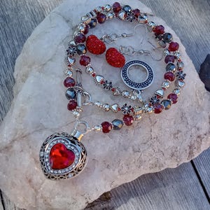 May include: A jewelry set featuring a necklace with a heart-shaped pendant, earrings, and a bracelet. The necklace has a silver toggle clasp and a red heart-shaped pendant with a crystal border. The set includes red and silver beads and flower accents.