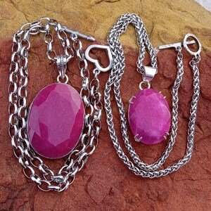 May include: Two silver necklaces with large, oval, magenta-colored gemstone pendants. The necklaces have different chain styles, one with a heart-shaped clasp and the other with a toggle clasp. The pendants are set in silver.