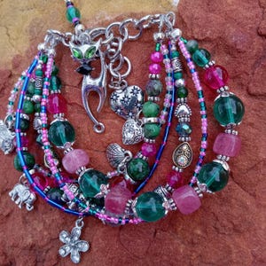 May include: A multi-strand bracelet featuring green, pink, and blue beads, accented with silver charms. Charms include a cat, hearts, a flower, and an elephant. The bracelet has a mix of round and square beads.