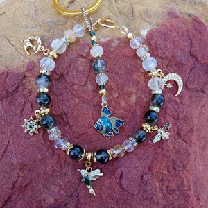 May include: A beaded bracelet with gold accents and charms. The bracelet features black, clear, and gold beads, with charms including a hummingbird, a fish, a star, a crescent moon, and a dragonfly. The bracelet is displayed on a textured, reddish-brown surface.