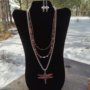 May include: A three-strand necklace set with matching earrings. The necklace features a silver chain, red and brown beads, and a dragonfly pendant. The earrings have elephant charms. The jewelry is displayed on a black mannequin.