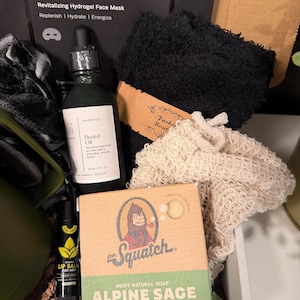 May include: A gift box containing men's grooming products. Includes a black face mask, beard oil, lip balm, a bar of Alpine Sage soap, and a black washcloth. The soap box features a cartoon Sasquatch.
