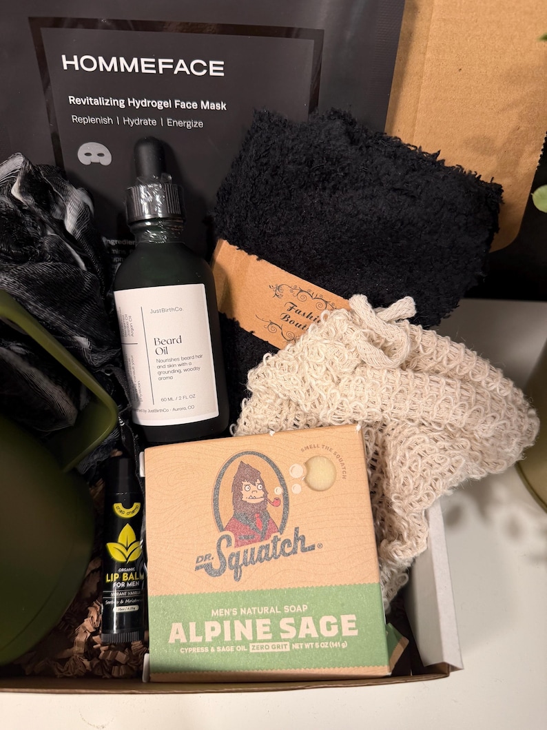 May include: A gift box containing men's grooming products. Includes a black face mask, beard oil, lip balm, a bar of Alpine Sage soap, and a black washcloth. The soap box features a cartoon Sasquatch.