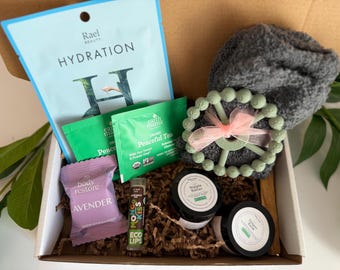 Può includere: Una scatola regalo contenente una maschera in tessuto azzurro Rael Hydration, due bustine di tè verde Earth Mama Peaceful Tea, un oggetto grigio soffice, un massaggiagengive circolare verde chiaro con un nastro rosa, una pastiglia doccia viola e due barattoli.