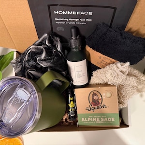 May include: An open gift box filled with grooming products. Includes a green tumbler, beard oil, a face mask, soap, a lip balm, and a black washcloth. The box is labeled "HOMMEFACE" and "Alpine Sage".
