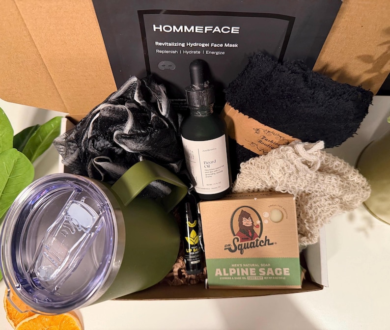 May include: An open gift box filled with grooming products. Includes a green tumbler, beard oil, a face mask, soap, a lip balm, and a black washcloth. The box is labeled "HOMMEFACE" and "Alpine Sage".