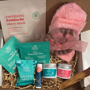 May include: A gift box filled with self-care items. Includes a pink plush baby sock with a ribbon, tea bags, a sheet mask, lip balm, diaper balm, and nipple butter. The box contains a variety of products for relaxation and skincare.