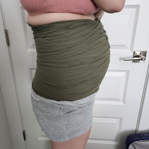 May include: An olive green belly band featuring a multi-layered, ruched fabric design with raw, distressed edges. The band is worn around the midsection, providing a textured, layered appearance that wraps horizontally across the torso.