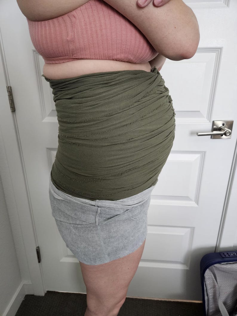 May include: An olive green belly band featuring a multi-layered, ruched fabric design with raw, distressed edges. The band is worn around the midsection, providing a textured, layered appearance that wraps horizontally across the torso.