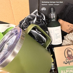 May include: An olive green travel mug with a handle, a bottle of beard oil, and a box of men's grooming products. The box includes a face mask, soap, and other items. The text "HOMMEFACE" is visible on the box.