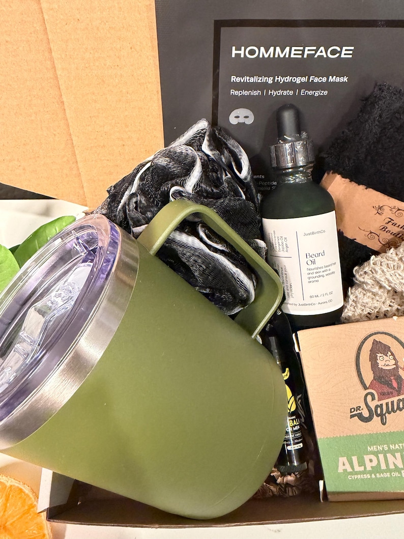 May include: An olive green travel mug with a handle, a bottle of beard oil, and a box of men's grooming products. The box includes a face mask, soap, and other items. The text "HOMMEFACE" is visible on the box.