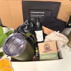 May include: An open cardboard box filled with self-care items. Includes a green travel mug, beard oil, face mask, soap, and other grooming products. The box contains the text "HOMMEFACE" and "Dr. Squatch ALPINE SAGE".