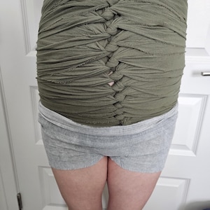 May include: An olive green fabric wrap featuring a series of horizontal strips gathered and knotted down the centre to create a textured, ruched appearance. The wrap is worn over a pair of light grey, heathered knit shorts with a wide elastic waistband.