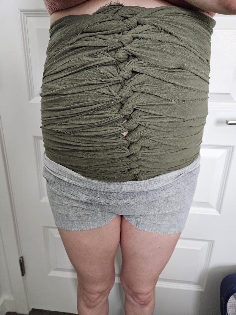 May include: An olive green fabric wrap featuring a series of horizontal strips gathered and knotted down the centre to create a textured, ruched appearance. The wrap is worn over a pair of light grey, heathered knit shorts with a wide elastic waistband.