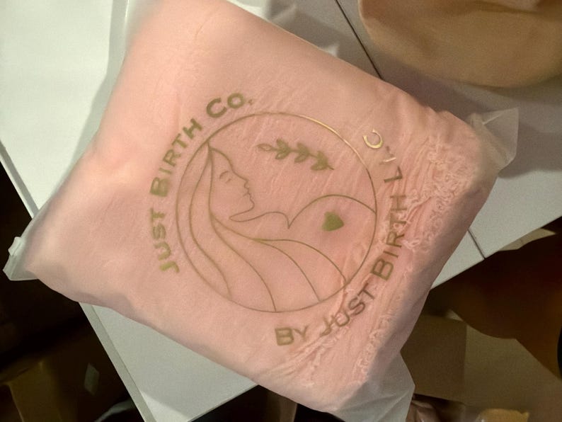 May include: A light pink square pouch made of a translucent material. The front features a gold circular logo with the text "JUST BIRTH CO." and "BY JUST BIRTH LLC" surrounding a line drawing of a person and a small leaf sprig.