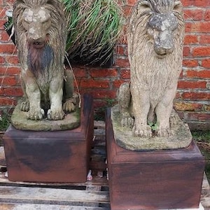 Op de afbeelding: Twee grijze stenen leeuwenbeelden op aparte bruine sokkels. De beelden staan op een houten pallet.