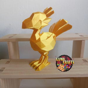 May include: A golden yellow, geometric-style figurine of a bird, possibly a Chocobo, is displayed on a wooden shelf. The bird has a polygonal design and is standing. The logo for Xamo Crafts is visible on the right.