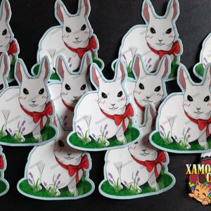 Puede incluir: Un conjunto de pegatinas de conejo blanco con lazos rojos. Cada pegatina presenta un conejo blanco con ojos rojos y un lazo rojo. Los conejos están de pie en un parche de hierba verde con flores moradas.