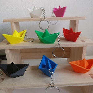 Puede incluir: Una colección de llaveros de barco origami de colores en blanco, rosa, amarillo, verde, rojo, negro, azul y naranja. Cada barco está unido a un llavero plateado. Los llaveros se exhiben en una pequeña estantería de madera.