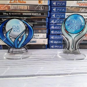 May include: Two blue and white PlayStation themed acrylic stands with a clear base. The stands feature a sphere with the PlayStation button symbols and a stylized design.
