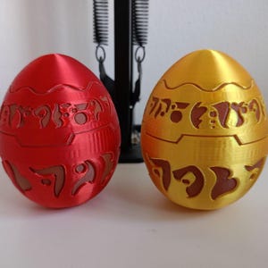 May include: Two decorative, egg-shaped objects. One is red, the other is gold. Both have intricate, carved designs and a two-part construction. The eggs are placed on a white surface, with a black object in the background.