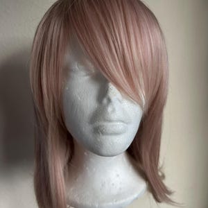 Puede incluir: Peluca rosa claro con cabello largo y en capas y un flequillo lateral. La peluca se exhibe en una cabeza de espuma de poliestireno blanca, mostrando el estilo y el color. El cabello tiene un aspecto natural, con variaciones sutiles.