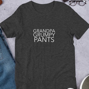 以下が含まれることがあります： 白で「GRANDPA GRUMPY PANTS」とプリントされた濃いグレーのTシャツです。