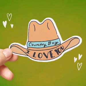 May include: A sticker featuring a cartoon cowboy hat in shades of tan and blue. The hat has the words "Country Boy I Love You" printed on it. The sticker is held by a hand against a green background with white heart doodles.