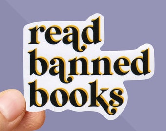 May include: A white sticker with the words "read banned books" in a black and yellow font. The sticker is held between two fingers against a purple background. The sticker is a rectangle with rounded edges.