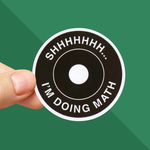 May include: A round black sticker with white text and a white circle in the center. The text reads "SHHHHHHHH..." at the top and "I'M DOING MATH" at the bottom. The sticker is held by a person's fingers against a green background.