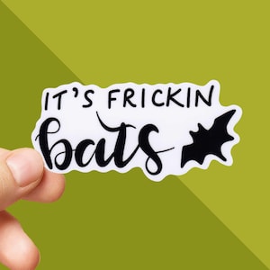 May include: A white sticker with the text "IT'S FRICKIN bats" in black script and a black bat silhouette. The sticker is held by a hand against a green background.