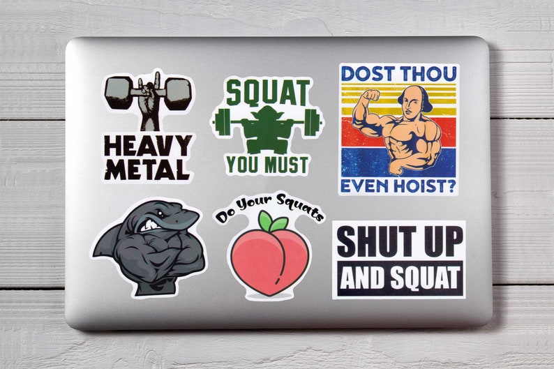 Op de afbeelding: Een verzameling fitness-thema stickers op een laptop. Ontwerpen omvatten een hand met gewichtheffer, een flexende haai, een perzik met de tekst "Do Your Squats" en teksten als "Heavy Metal", "Squat You Must" en "Shut Up and Squat".