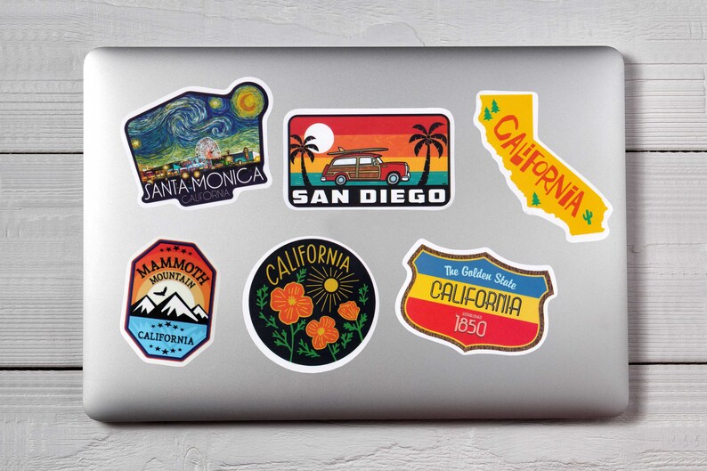 May include: A collection of colorful California-themed stickers on a laptop. Designs include Santa Monica, San Diego, Mammoth Mountain, and the state outline, featuring vibrant illustrations and text.