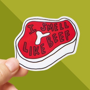 May include: A sticker in the shape of a steak, with the words "I SMELL LIKE BEEF" in red letters. The steak is red with a white bone-shaped design in the middle, outlined in black. The sticker is held against a green background.