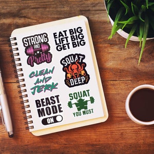 Op de afbeelding: Een spiraalgebonden notitieboek met fitness-thema stickers op een houten oppervlak. De stickers tonen zinnen als "Strong and Pretty", "Eat Big, Lift Big, Get Big" en "Squat Deep". Een pen, een kop koffie en een plant zijn ook aanwezig.