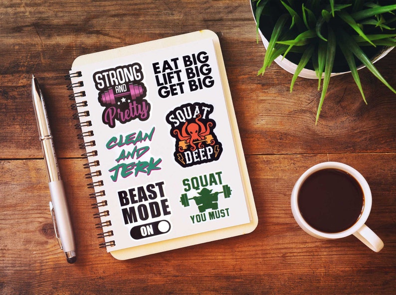 Op de afbeelding: Een spiraalgebonden notitieboek met fitness-thema stickers op een houten oppervlak. De stickers tonen zinnen als "Strong and Pretty", "Eat Big, Lift Big, Get Big" en "Squat Deep". Een pen, een kop koffie en een plant zijn ook aanwezig.