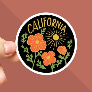 Peut inclure: Un autocollant rond avec un fond noir. Il pr&eacute;sente le mot "CALIFORNIA" en jaune, un soleil et des fleurs de pavot orange. L'autocollant a une bordure blanche et est tenu par une main.