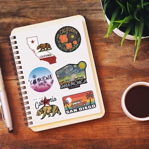 May include: A spiral-bound notebook with several California-themed stickers. The stickers feature images of the California state outline, a bear, palm trees, and the text "California", "Santa Monica", and "San Diego". A pen, a cup of coffee, and a plant are also on the wooden surface.