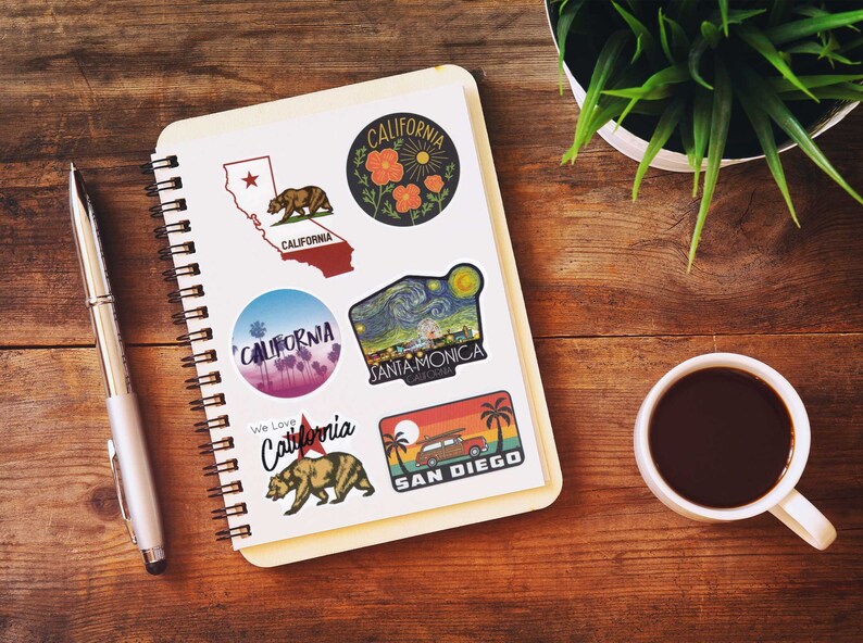 May include: A spiral-bound notebook with several California-themed stickers. The stickers feature images of the California state outline, a bear, palm trees, and the text "California", "Santa Monica", and "San Diego". A pen, a cup of coffee, and a plant are also on the wooden surface.