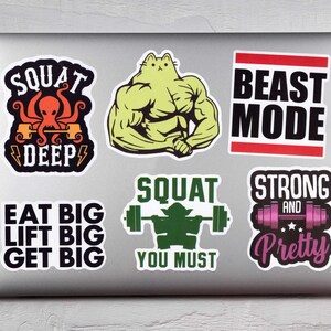 Op de afbeelding: Een verzameling fitness-thema stickers op een laptop. De stickers tonen slogans als "SQUAT DEEP", "BEAST MODE" en "EAT BIG LIFT BIG GET BIG". Andere stickers tonen een gespierde kat en een Yoda-achtig figuur dat gewichten heft.