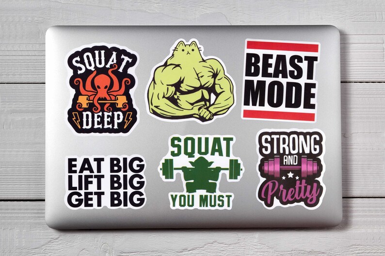 Op de afbeelding: Een verzameling fitness-thema stickers op een laptop. De stickers tonen slogans als "SQUAT DEEP", "BEAST MODE" en "EAT BIG LIFT BIG GET BIG". Andere stickers tonen een gespierde kat en een Yoda-achtig figuur dat gewichten heft.