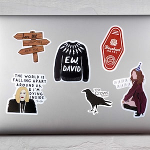May include: A collection of stickers on a laptop. The stickers include a wooden sign, a black sweater with "EW, DAVID" text, a red motel key, a woman with text, a crow, and a woman with party hat.